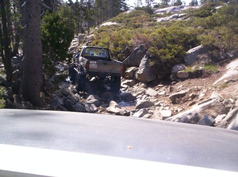 1989 Ford Bronco - A pickup truck drives through a rocky stream in a wooded, mountainous area.