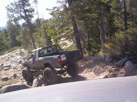 1989 Ford Bronco - A lifted pickup truck maneuvers through a challenging, rocky off-road course surrounded by trees.