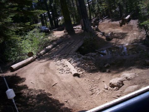 1989 Ford Bronco - A dark SUV navigates a challenging dirt trail with obstacles like logs and rocks.