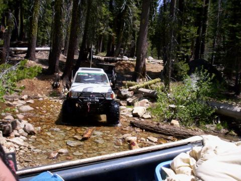 1989 Ford Bronco - White off-road SUV navigates a rocky stream bed surrounded by dense forest.