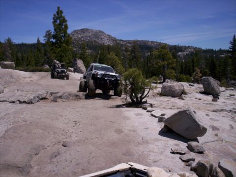 1989 Ford Bronco - Two off-road vehicles traverse a challenging rocky trail in a mountainous, wooded area on a sunny day.