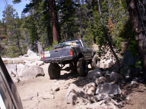 1989 Ford Bronco - This modified pickup truck is shown off-roading on a challenging, rocky path surrounded by trees.
