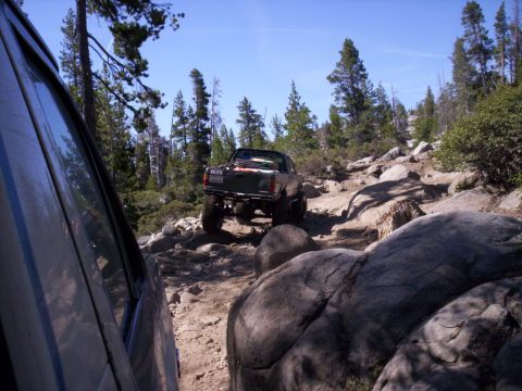 1989 Ford Bronco - Two vehicles navigate a challenging, boulder-strewn path through a forest on a sunny day.