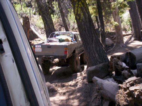 1989 Ford Bronco - A lifted pickup truck navigates a narrow, wooded trail, with a portion of another vehicle visible in the foreground.