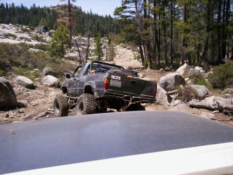 1989 Ford Bronco - Pickup truck navigating a rough, rocky off-road trail with trees and boulders.