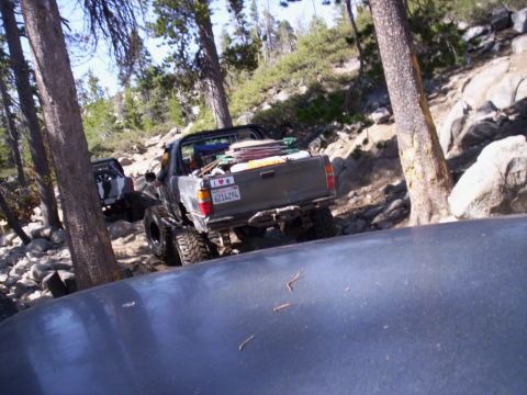 1989 Ford Bronco - Two modified pickup trucks navigate a challenging, rocky off-road trail through a wooded area.