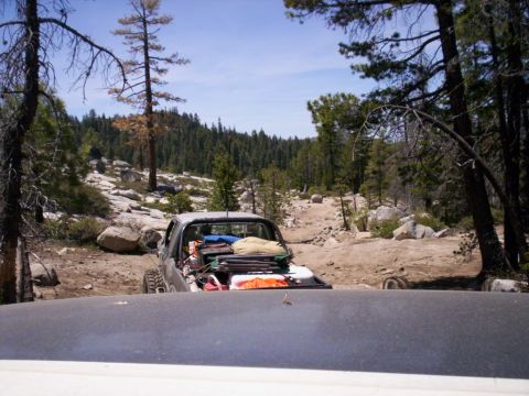 1989 Ford Bronco - An off-road vehicle packed with gear navigates a rocky trail through a wooded area.