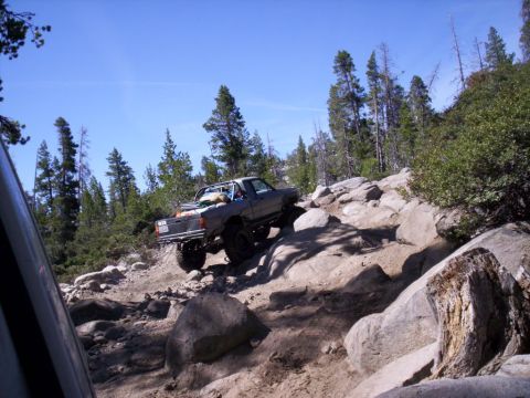 1989 Ford Bronco - A modified pickup truck is shown off-roading on a steep, rocky incline with trees in the background.