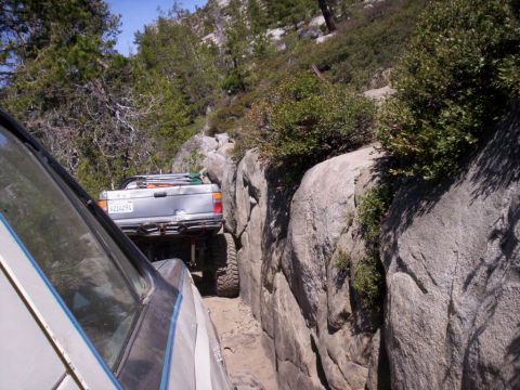 1989 Ford Bronco - Two vehicles are carefully maneuvering through a narrow, rocky mountain pass with trees and brush on either side.