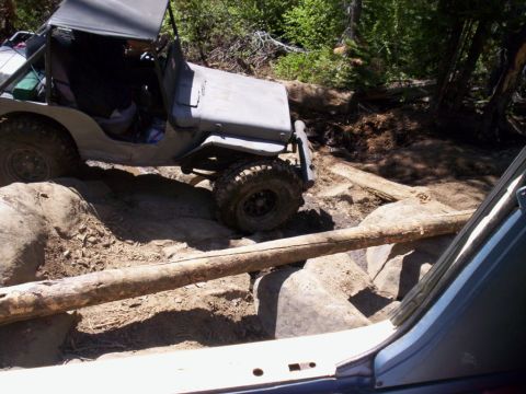 1989 Ford Bronco - An off-road vehicle traverses a challenging trail with large rocks and fallen logs in a wooded area.