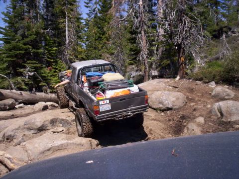 1989 Ford Bronco - Pickup truck loaded with gear navigating a challenging off-road trail through a forest.