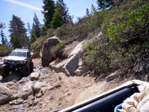 1989 Ford Bronco - An off-road vehicle navigates a challenging rocky path with dense vegetation and large boulders surrounding the trail.