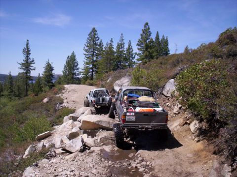 1989 Ford Bronco - Two off-road vehicles navigate a challenging rocky path through a wooded, mountainous area.
