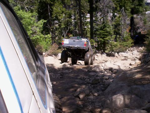 1989 Ford Bronco - Two off-road vehicles traverse a challenging, rocky path through a wooded area.