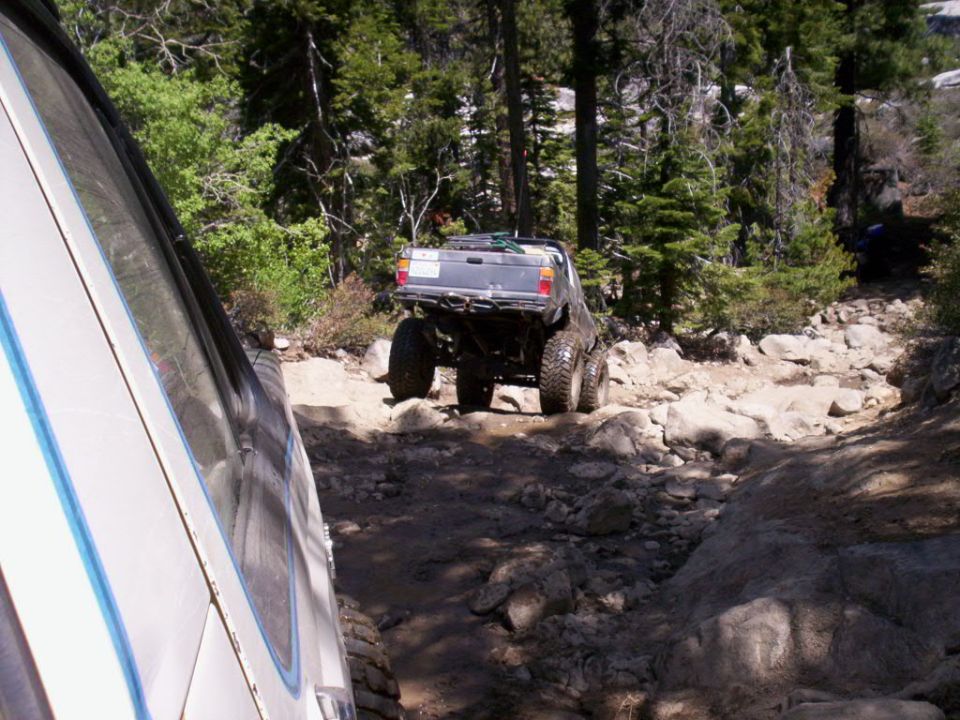 Gray off-road truck with large tires navigating a rocky trail, viewed from behind and to the side