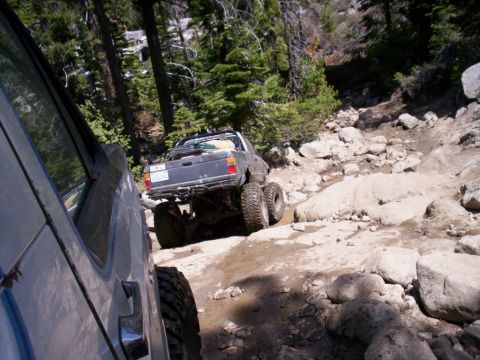 1989 Ford Bronco - A heavily modified pickup truck with dual rear wheels tackles a challenging rocky off-road trail, with another vehicle visible in the foreg…