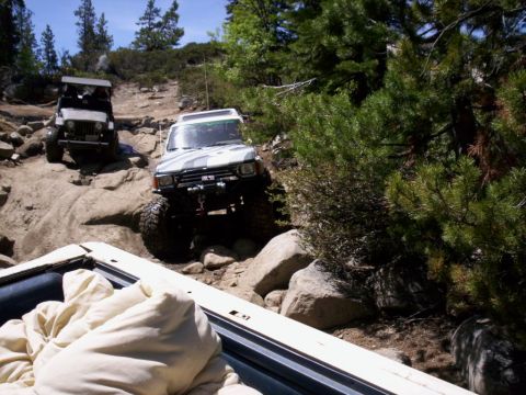 1989 Ford Bronco - Two modified off-road vehicles navigate a challenging rocky trail in a wooded area.