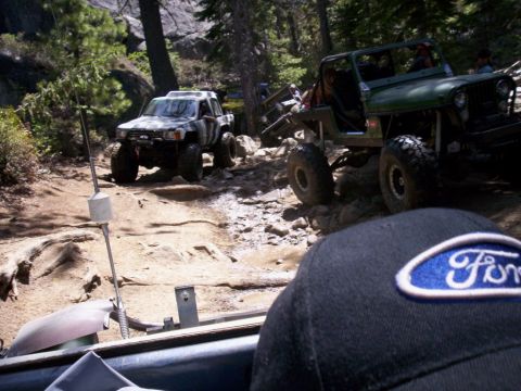1989 Ford Bronco - Multiple off-road vehicles traverse a challenging, rocky path through a wooded area.