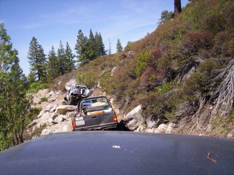 1989 Ford Bronco - Two off-road vehicles carefully make their way up a challenging, boulder-strewn mountain path under a clear blue sky.