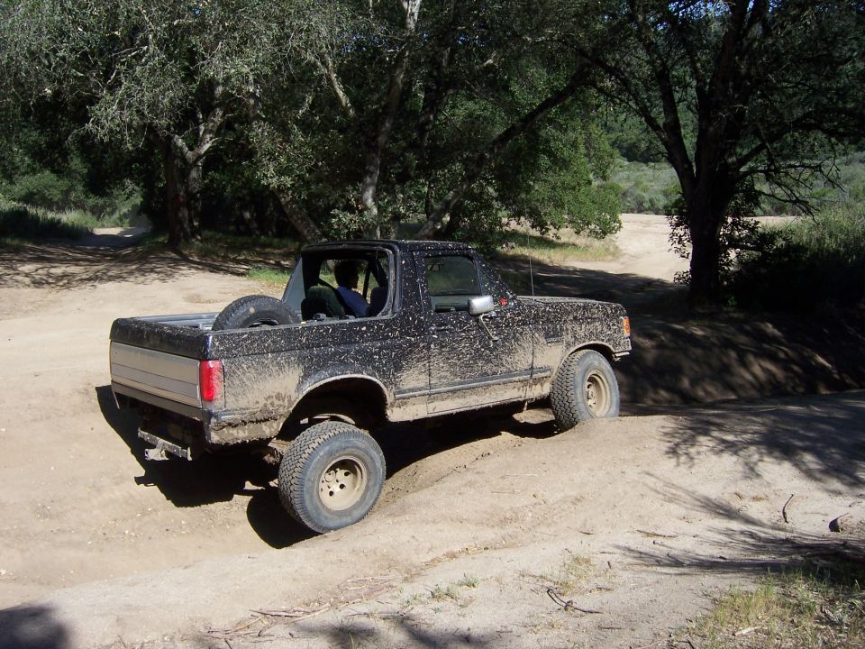 Mud-covered 1991 Ford Bronco driving on a dirt trail with trees in the background