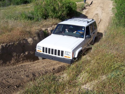 1991 Ford Bronco - A white Jeep Cherokee XJ navigates a challenging off-road trail with steep inclines and rough terrain.