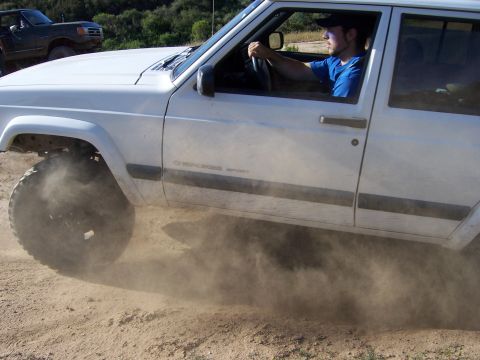 1991 Ford Bronco - A white Jeep Cherokee Sport drives off-road, kicking up a cloud of dust.