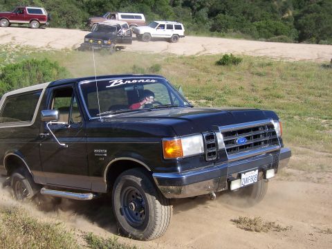 1991 Ford Bronco - A black 1991 Ford Bronco kicks up dust as it drives on a dirt road, with several other Broncos visible in the background.