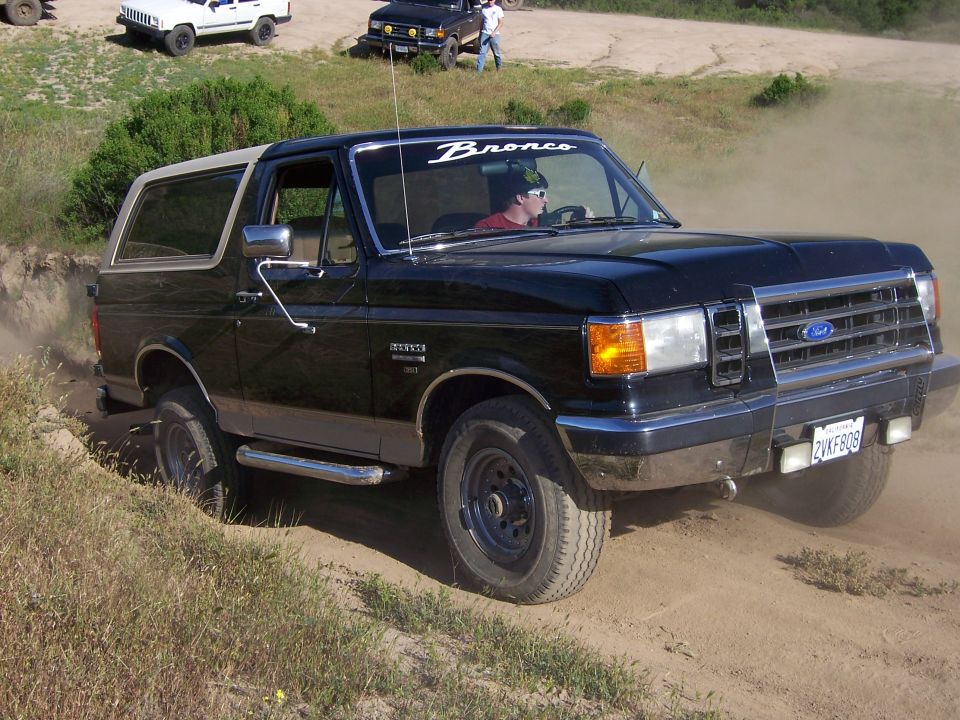 Black 1991 Ford Bronco driving off-road, kicking up dust. White Jeep and black SUV in background