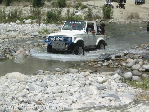 1987 Suzuki Samurai - A white Suzuki Samurai navigates a shallow, rocky riverbed, kicking up water as it goes.