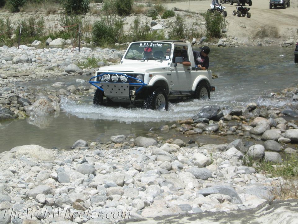 White 1987 Suzuki Samurai with blue bumper and off-road lights driving through a rocky stream. R.F.L. Racing sticker on windshield. Other ATVs and vehicles in background