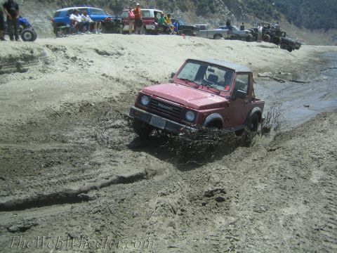 1987 Suzuki Samurai - Red Suzuki Samurai navigating a muddy trail with other vehicles and spectators nearby.