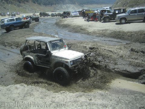 1987 Suzuki Samurai - A white Suzuki Samurai navigates a challenging muddy terrain, sending mud flying.