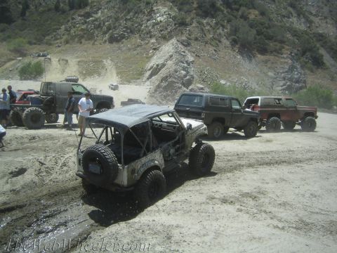 1987 Suzuki Samurai - Multiple off-road vehicles navigate a muddy and dusty terrain with steep hills in the background.