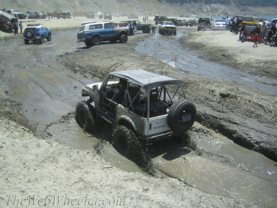 White Suzuki Samurai off-roading in mud, rear view. Modified with roll cage and large tires