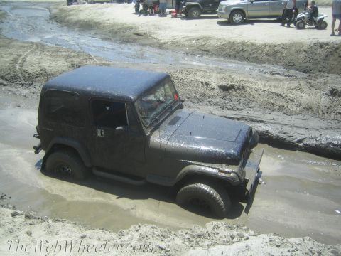 1987 Suzuki Samurai - A dark Jeep Wrangler is stuck in a muddy track with other vehicles and people in the background.