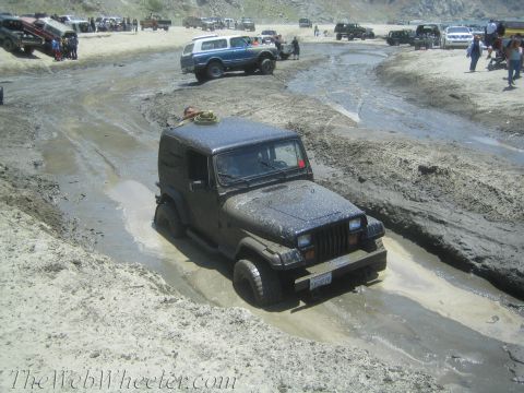 1987 Suzuki Samurai - A dark Jeep Wrangler struggles through a muddy off-road course, with several other vehicles and spectators visible in the distance.