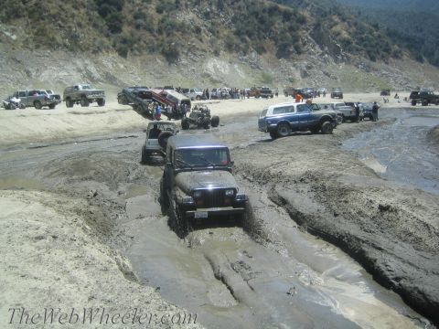 1987 Suzuki Samurai - Multiple off-road vehicles navigate a challenging muddy terrain, with spectators gathered in the background.