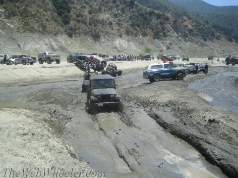 1987 Suzuki Samurai - Multiple off-road vehicles are seen driving through a muddy area, with a crowd gathered in the distance.