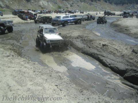 1987 Suzuki Samurai - Off-road vehicles navigate a muddy track under a rocky hillside.