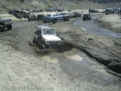 1987 Suzuki Samurai - A white 1987 Suzuki Samurai navigates a muddy track, kicking up water and dirt, with a crowd of off-road vehicles gathered nearby.