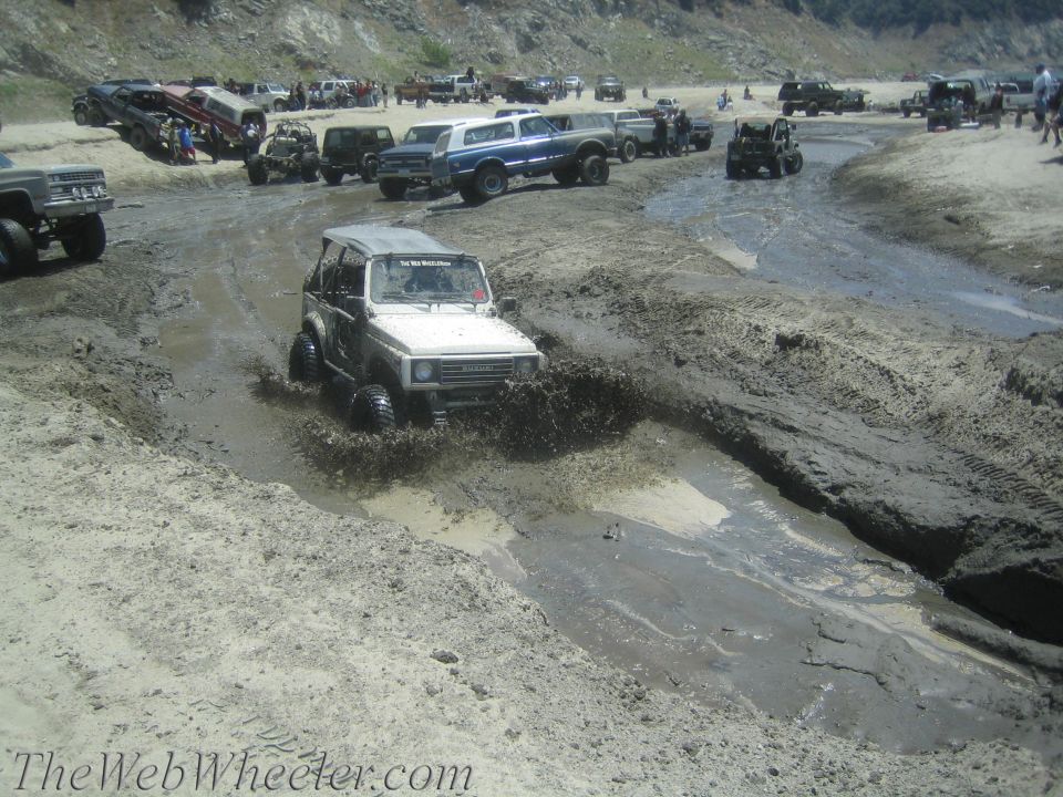 White 1987 Suzuki Samurai splashing through mud with other off-road vehicles in background