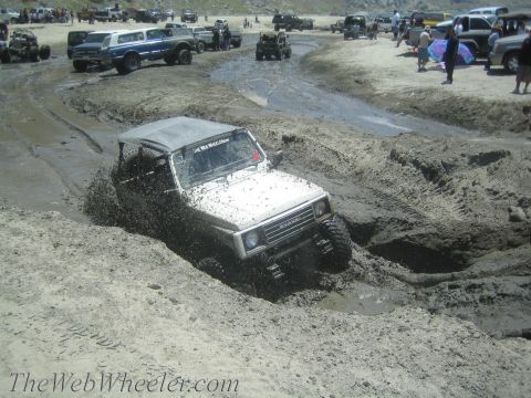 1987 Suzuki Samurai - A white 1987 Suzuki Samurai tackles a challenging muddy terrain, with other vehicles and spectators visible in the background.