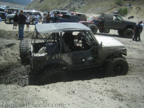 1987 Suzuki Samurai - A muddy 1987 Suzuki Samurai navigates rough terrain, with other vehicles and people in the background.