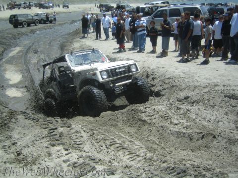 1987 Suzuki Samurai - A white 1987 Suzuki Samurai navigates a challenging muddy obstacle on a beach with spectators watching.
