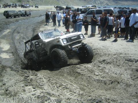 1987 Suzuki Samurai - A white 1987 Suzuki Samurai tackles a challenging muddy terrain, with a crowd observing the off-road action.