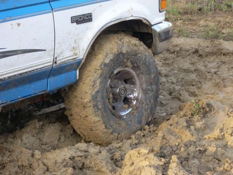 1992 Ford Bronco - A white and blue 1992 Ford Bronco is stuck in thick mud, with its front tire heavily caked in dirt.