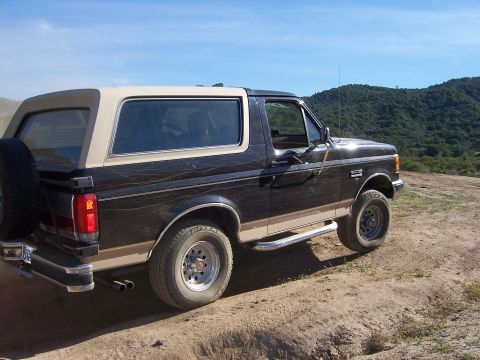 1991 Ford Bronco - A dark-colored Ford Bronco with a tan top is parked on a dirt path, with rolling green hills under a blue sky.