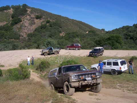 1991 Ford Bronco - Several off-road vehicles are gathered on a dusty trail with a hilly, wooded landscape under a clear blue sky.