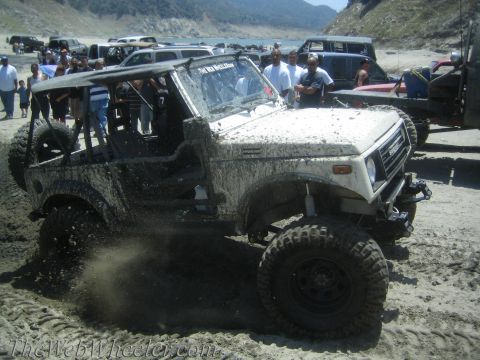 1987 Suzuki Samurai - A white 1987 Suzuki Samurai is stuck in the mud, with other vehicles and people in the background near a body of water.