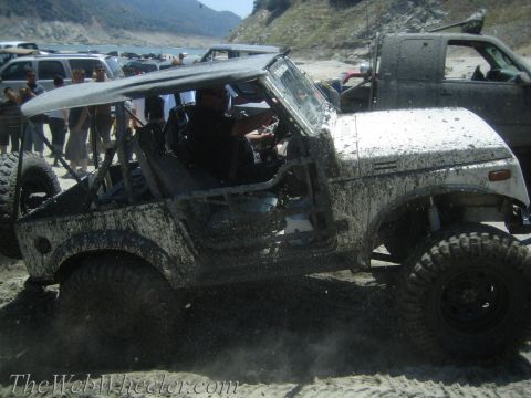 1987 Suzuki Samurai - Off-road Suzuki Samurai covered in mud, kicking up sand near a lake with other vehicles present.